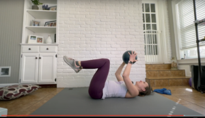 Fitness trainer lying on her back with knees over her hips and a pilates ball in her arms extended over her shoulders.