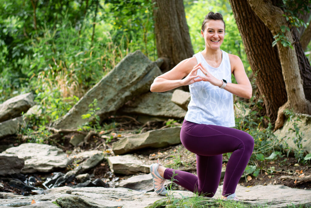 A female fitness trainer is doing a lunge and twisting on a rock along a river. She's wearing a white tank top and purple leggings.