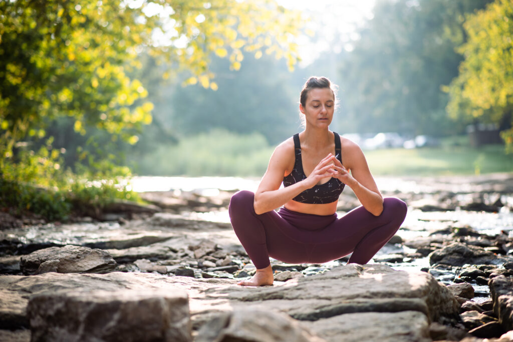 Female fitness trainer sitting in a deep squat on a rock on a river. Her eyes are closed as if she's in deep thought. She's wearing a black sports bra and purple leggings.